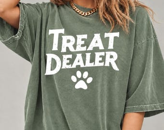 May include: An olive green short-sleeved t-shirt with a distressed white graphic print on the chest that reads "TREAT DEALER" in bold, capitalized letters above a simple white paw print icon.