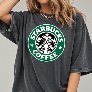 May include: A charcoal gray short-sleeve t-shirt featuring a large, circular green and white Starbucks Coffee logo printed on the center of the chest. The garment has a relaxed, oversized fit with a crew neckline and a washed-out fabric texture.