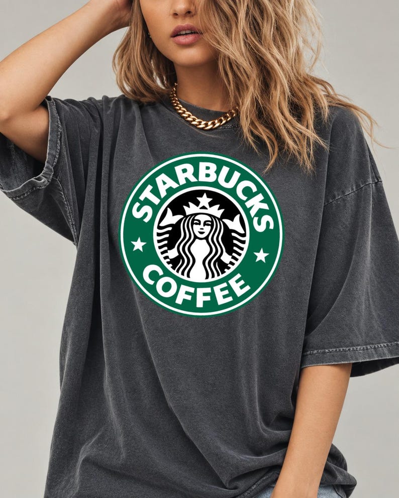 May include: A charcoal gray short-sleeve t-shirt featuring a large, circular green and white Starbucks Coffee logo printed on the center of the chest. The garment has a relaxed, oversized fit with a crew neckline and a washed-out fabric texture.