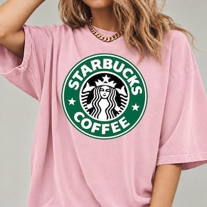 May include: A light pink short-sleeved t-shirt featuring a large, circular green and white Starbucks Coffee logo printed on the center of the chest. The shirt has a relaxed, loose fit with a crew neckline and a solid, muted pink color.