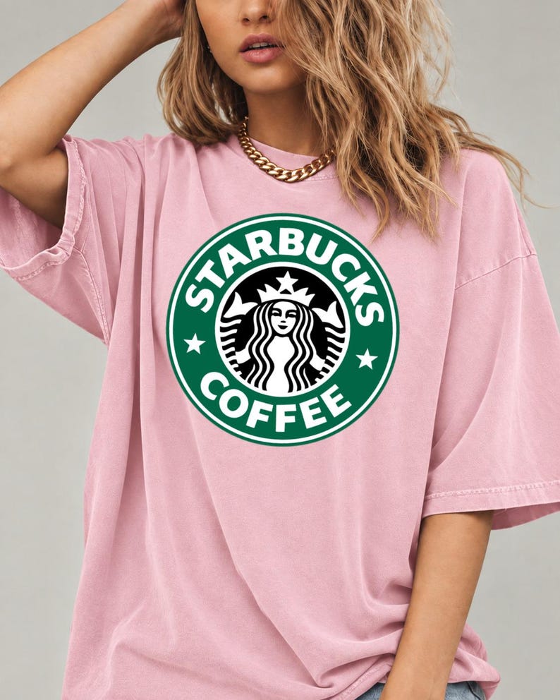 May include: A light pink short-sleeved t-shirt featuring a large, circular green and white Starbucks Coffee logo printed on the center of the chest. The shirt has a relaxed, loose fit with a crew neckline and a solid, muted pink color.