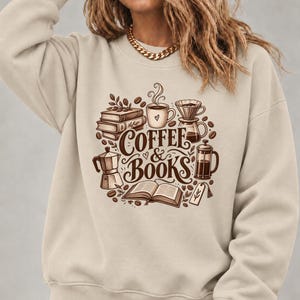 May include: A beige crewneck sweatshirt featuring a brown graphic print of stacked books, an open book, a coffee mug, a French press, a moka pot, and a pour-over dripper. The text "COFFEE & BOOKS" is centered in the middle of the illustration.