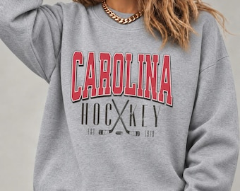 May include: A heather gray crewneck sweatshirt featuring the red text CAROLINA above the black text HOCKEY with a graphic of two crossed hockey sticks and a puck. The text EST 1979 is printed on either side of the hockey sticks.