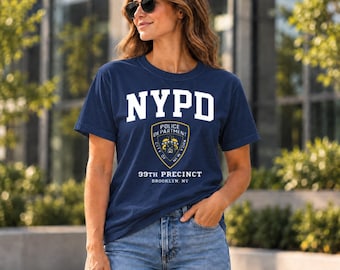 May include: A navy blue short-sleeved t-shirt featuring the white text NYPD above a police department shield emblem. Below the emblem, the text reads 99TH PRECINCT BROOKLYN, NY in white lettering. The shirt is worn with blue denim jeans.