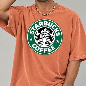 May include: A short-sleeved, crew-neck t-shirt in a washed-out terracotta orange color. The center of the chest features a large, circular green and white Starbucks Coffee logo with the iconic siren graphic and text printed clearly.