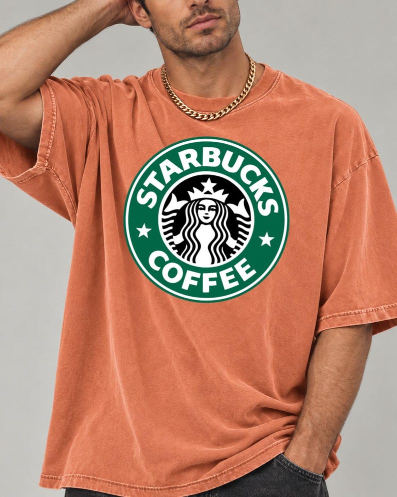 May include: A short-sleeved, crew-neck t-shirt in a washed-out terracotta orange color. The center of the chest features a large, circular green and white Starbucks Coffee logo with the iconic siren graphic and text printed clearly.