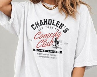 May include: A white short-sleeved t-shirt featuring a graphic that reads Chandler's New York City Comedy Club Estd 1994, The One With The Jokes, Where Sarcasm Thrives & Awkwardness Survives, with an illustration of a microphone.
