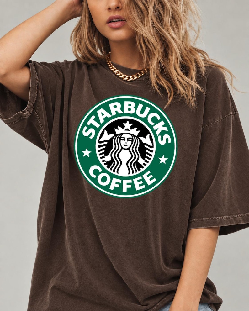 May include: A dark brown, short-sleeved t-shirt featuring the green and white Starbucks Coffee circular logo printed on the center of the chest. The garment has a relaxed, loose fit with a crew neckline and a slightly faded, vintage-style fabric texture.