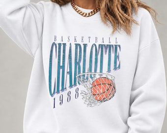 May include: A white long-sleeve crewneck sweatshirt featuring a graphic print with the text "BASKETBALL CHARLOTTE 1988" in blue and purple, accompanied by an illustration of an orange basketball going through a white net.