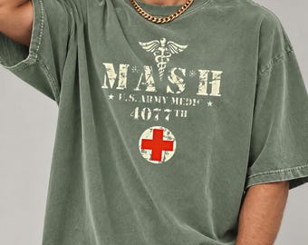 May include: An olive green short-sleeve t-shirt featuring a distressed graphic with the text MASH, U.S. ARMY MEDIC, and 4077TH, accompanied by a caduceus symbol and a white circle containing a red cross emblem on the chest.
