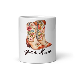 May include: A white ceramic mug featuring a graphic of two cream-colored cowboy boots decorated with colorful floral patterns in shades of red, yellow, and blue. Below the boots, the text yee haw is written in a dark brown cursive script.