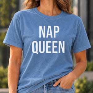 Könnte beinhalten: Ein blaues kurzärmeliges T-Shirt mit Rundhalsausschnitt und dem Text NAP QUEEN in fetten, weißen Großbuchstaben auf der Brust. Das Kleidungsstück besteht aus einem weichen, melierten Stoff und wird mit einer blauen Jeans kombiniert.