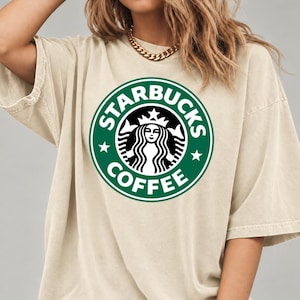 May include: A beige short-sleeved t-shirt featuring a large, circular green and white Starbucks Coffee logo printed on the center of the chest. The garment has a crew neckline and a relaxed, loose-fitting silhouette with dropped shoulder seams.