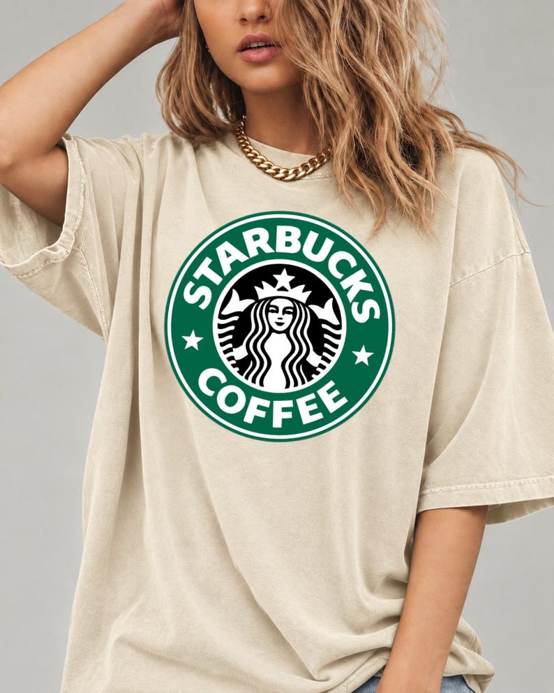 May include: A beige short-sleeved t-shirt featuring a large, circular green and white Starbucks Coffee logo printed on the center of the chest. The garment has a crew neckline and a relaxed, loose-fitting silhouette with dropped shoulder seams.