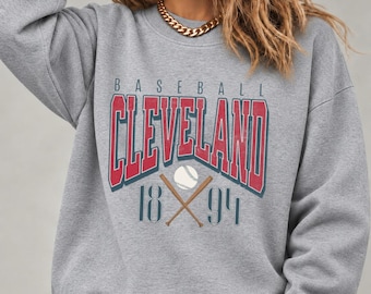 May include: A heather gray crewneck sweatshirt featuring a graphic print with the text BASEBALL CLEVELAND 1894. The design includes red block lettering, a white baseball, and two crossed brown wooden bats centered on the chest.