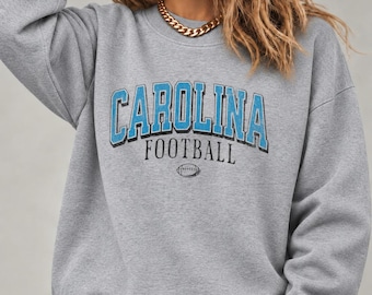 May include: A heather gray crewneck sweatshirt featuring the text CAROLINA in large blue letters with a black outline, the word FOOTBALL in smaller black letters, and a small graphic of a football centered below the text.