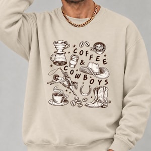May include: A beige crewneck sweatshirt featuring a brown graphic print of coffee-related items and western motifs, including a cowboy on a bucking horse, a hat, boots, and the text "COFFEE & COWBOYS" with small star accents.