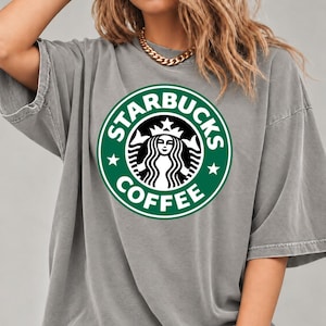 May include: A grey short-sleeved t-shirt featuring a large, circular green and white Starbucks Coffee logo graphic on the chest. The shirt has a relaxed, loose fit with a crew neckline and a washed, slightly faded fabric appearance.