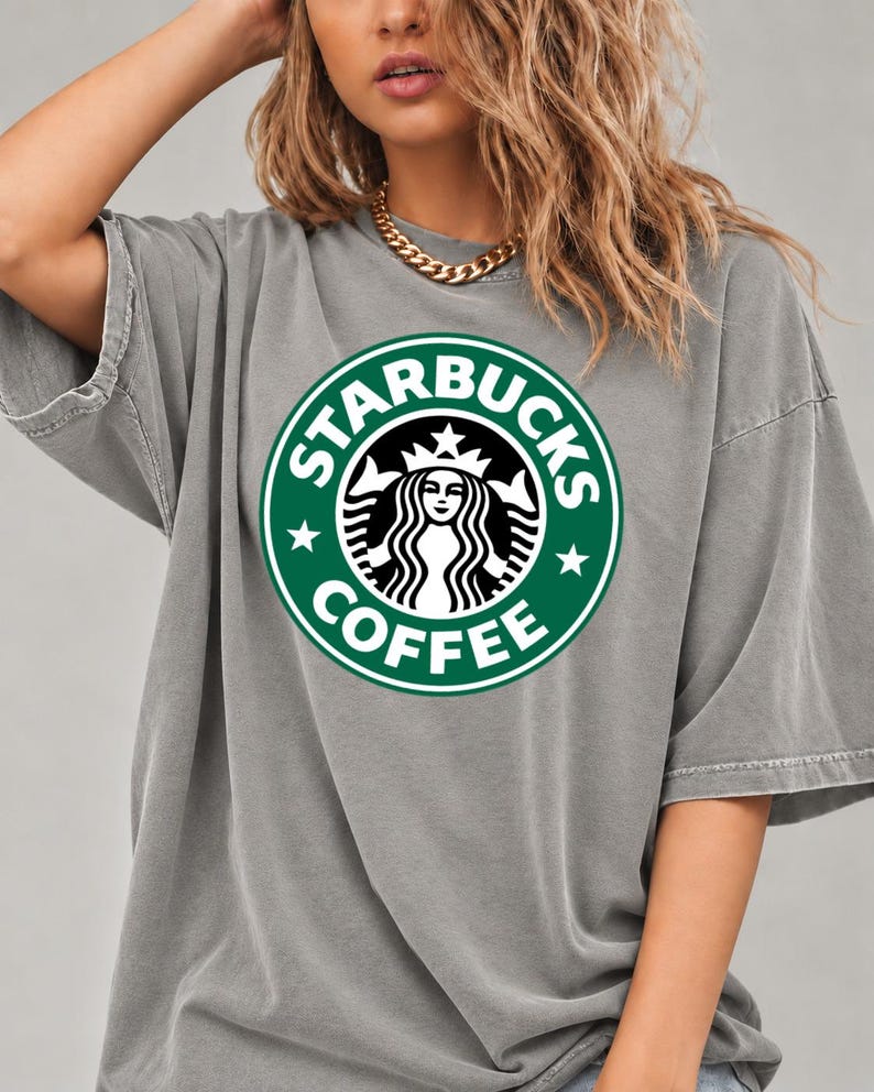 May include: A grey short-sleeved t-shirt featuring a large, circular green and white Starbucks Coffee logo graphic on the chest. The shirt has a relaxed, loose fit with a crew neckline and a washed, slightly faded fabric appearance.