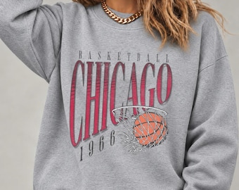 May include: A grey crewneck sweatshirt featuring a graphic print with the text "BASKETBALL CHICAGO 1966" in red and black lettering, accompanied by an illustration of an orange basketball passing through a white net.