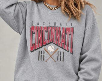 May include: A heather gray crewneck sweatshirt featuring a graphic print with the text BASEBALL CINCINNATI 1881. The design includes red block lettering, a white baseball, and two crossed wooden bats centered on the chest.