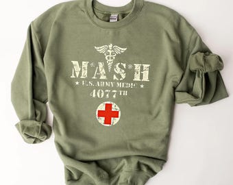 May include: An olive green crewneck sweatshirt featuring a distressed white graphic that reads MASH U.S. ARMY MEDIC 4077TH with a caduceus symbol and a circular red cross emblem centered on the chest area of the garment.