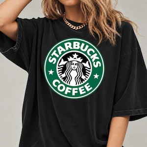 May include: A black short-sleeved t-shirt featuring a large, circular green and white Starbucks Coffee logo printed on the chest. The logo depicts a stylized siren figure surrounded by the text Starbucks Coffee and two small stars.