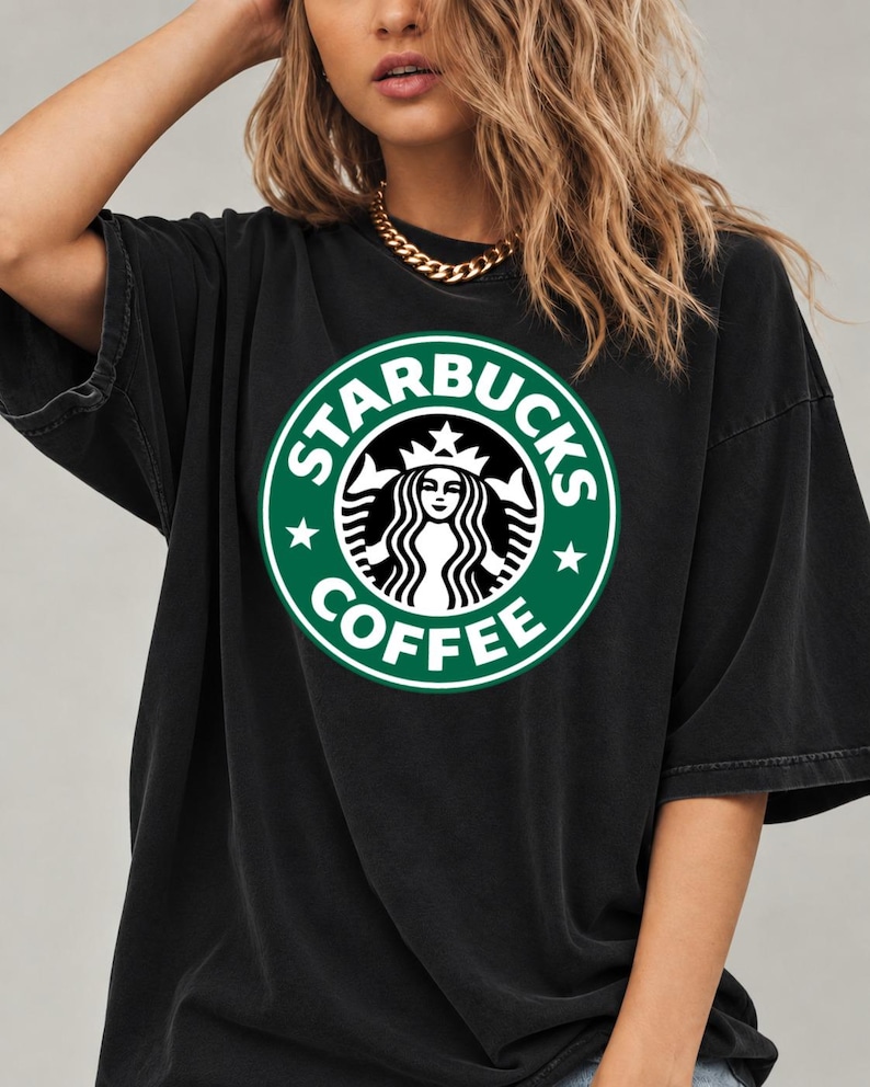 May include: A black short-sleeved t-shirt featuring a large, circular green and white Starbucks Coffee logo printed on the chest. The logo depicts a stylized siren figure surrounded by the text Starbucks Coffee and two small stars.