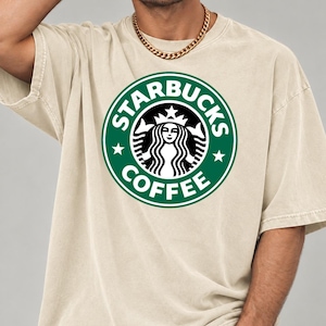 May include: A beige short-sleeve t-shirt featuring a large, circular green and white Starbucks Coffee logo printed on the chest. The graphic displays the iconic siren figure surrounded by the text Starbucks Coffee and two small stars.