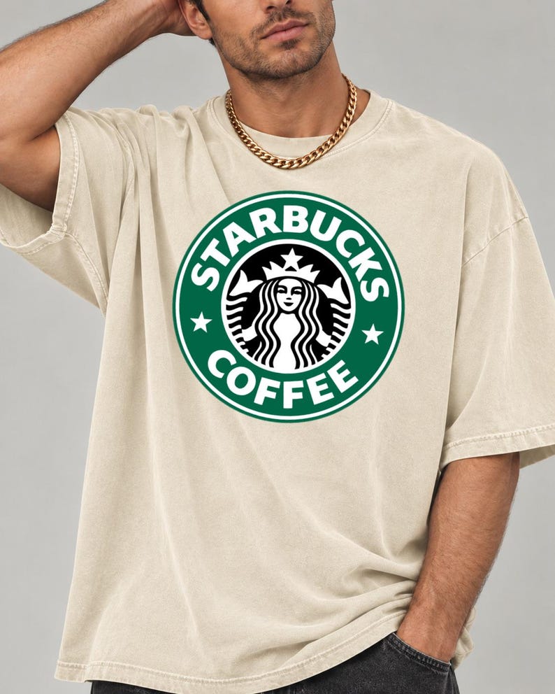 May include: A beige short-sleeve t-shirt featuring a large, circular green and white Starbucks Coffee logo printed on the chest. The graphic displays the iconic siren figure surrounded by the text Starbucks Coffee and two small stars.