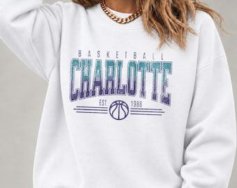 May include: A white long-sleeve sweatshirt featuring a graphic print that reads "BASKETBALL CHARLOTTE EST. 1988" in a distressed teal and purple gradient font, accompanied by a central basketball icon and horizontal lines.