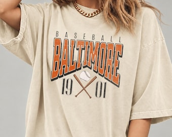 May include: A beige short-sleeve t-shirt featuring the text BASEBALL BALTIMORE 1901 in orange and black lettering. The graphic includes an illustration of a white baseball and two crossed brown wooden baseball bats centered below the city name.