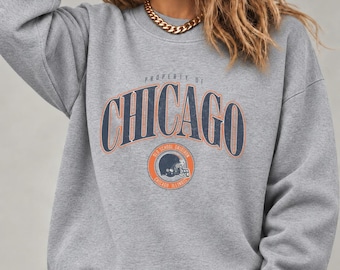 May include: A heather gray crewneck sweatshirt featuring the text Property of Chicago in dark blue and orange lettering. Below the text is a circular graphic depicting a football helmet with the words Old School Gridiron Chicago Illinois.