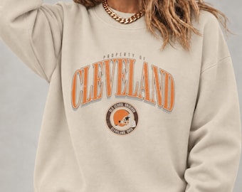 May include: A beige crewneck sweatshirt featuring the text PROPERTY OF CLEVELAND in an arched orange font with a white outline. Below the text is a circular logo with a football helmet graphic and the words OLD SCHOOL GRIDIRON CLEVELAND OHIO.