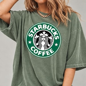 May include: An olive green short-sleeve t-shirt featuring a large, circular Starbucks Coffee logo printed on the chest. The logo displays the iconic siren graphic in white against a dark green background with the text STARBUCKS COFFEE.