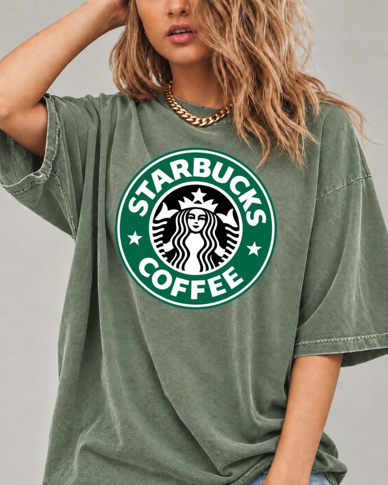 May include: An olive green short-sleeve t-shirt featuring a large, circular Starbucks Coffee logo printed on the chest. The logo displays the iconic siren graphic in white against a dark green background with the text STARBUCKS COFFEE.