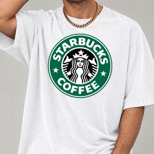 May include: A white short-sleeved t-shirt featuring the green and white Starbucks Coffee circular logo printed on the chest. The graphic includes the iconic siren figure, stars, and the text Starbucks Coffee in a bold, circular design.