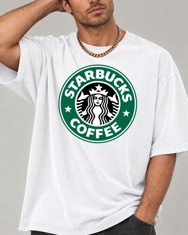 May include: A white short-sleeved t-shirt featuring the green and white Starbucks Coffee circular logo printed on the chest. The graphic includes the iconic siren figure, stars, and the text Starbucks Coffee in a bold, circular design.
