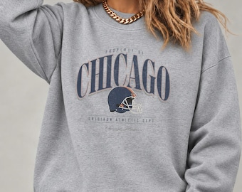 May include: A heather gray crewneck sweatshirt featuring the text Property of Chicago in dark blue arched lettering. Below the text is a graphic of a dark blue football helmet with a white facemask and the text Gridiron Athletic Dept.