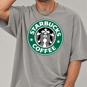 May include: A grey short-sleeved t-shirt featuring a large, circular green and white Starbucks Coffee logo printed on the chest. The logo displays the iconic siren graphic, the text STARBUCKS COFFEE, and two small stars on the sides.