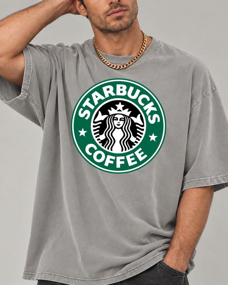 May include: A grey short-sleeved t-shirt featuring a large, circular green and white Starbucks Coffee logo printed on the chest. The logo displays the iconic siren graphic, the text STARBUCKS COFFEE, and two small stars on the sides.
