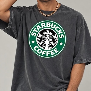 May include: A charcoal gray short-sleeve t-shirt featuring a large, circular green and white Starbucks Coffee logo printed on the chest. The shirt has a crew neckline and a relaxed, loose-fitting silhouette with visible seam detailing.