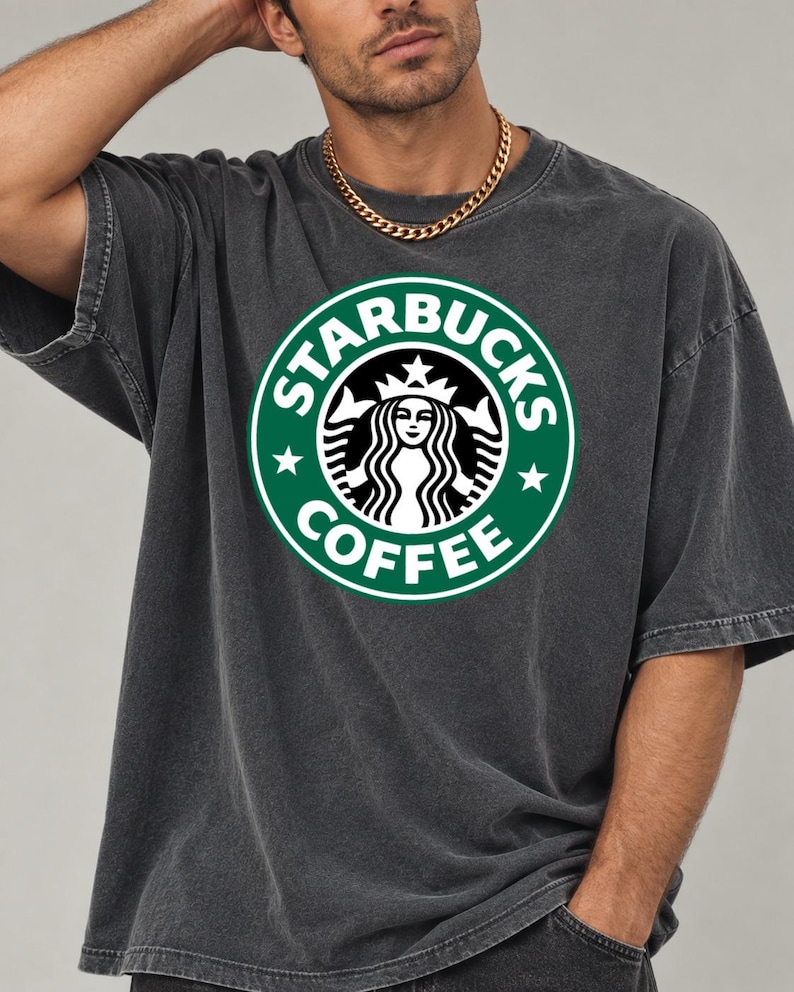 May include: A charcoal gray short-sleeve t-shirt featuring a large, circular green and white Starbucks Coffee logo printed on the chest. The shirt has a crew neckline and a relaxed, loose-fitting silhouette with visible seam detailing.
