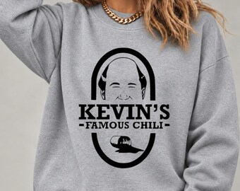 May include: A heather grey crewneck sweatshirt featuring a black graphic print. The design includes a stylised illustration of a man's face inside an oval, with the text KEVIN'S FAMOUS CHILI and a small spilled pot icon below the lettering.