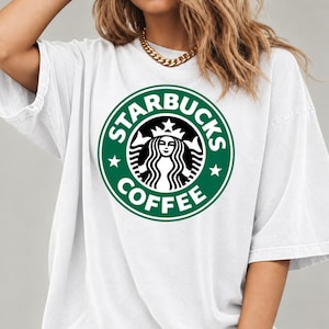 May include: A white short-sleeved t-shirt featuring a large, circular green and white Starbucks Coffee logo graphic printed on the center of the chest. The design includes the iconic siren figure surrounded by the text Starbucks Coffee.
