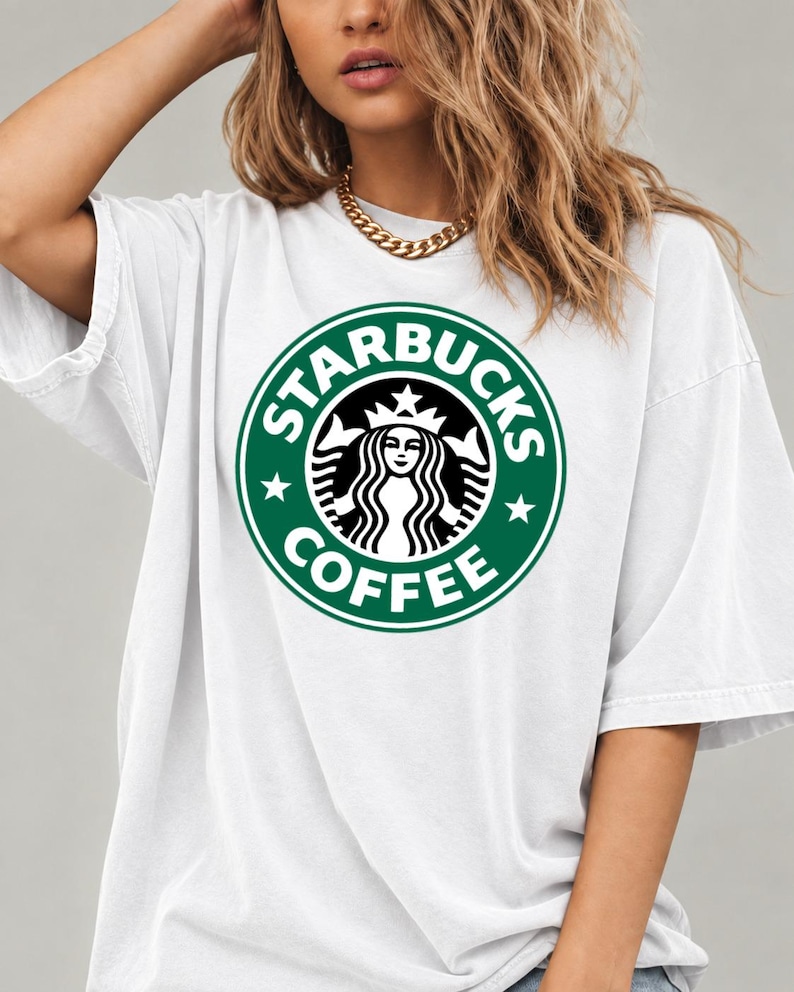 May include: A white short-sleeved t-shirt featuring a large, circular green and white Starbucks Coffee logo graphic printed on the center of the chest. The design includes the iconic siren figure surrounded by the text Starbucks Coffee.