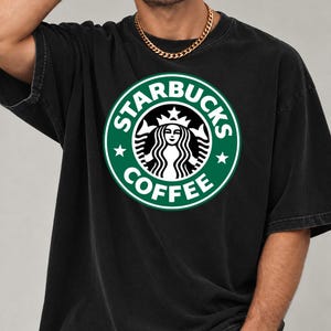 May include: A black short-sleeved t-shirt featuring a large, circular green and white Starbucks Coffee logo graphic on the chest. The logo depicts a stylized siren figure with a crown, surrounded by the text Starbucks Coffee and two stars.