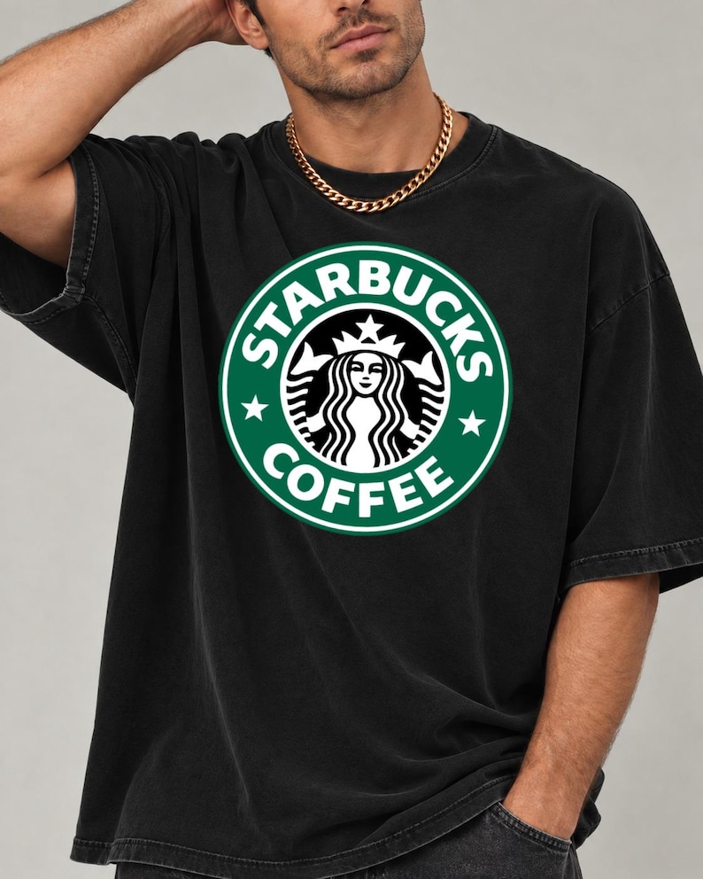 May include: A black short-sleeved t-shirt featuring a large, circular green and white Starbucks Coffee logo graphic on the chest. The logo depicts a stylized siren figure with a crown, surrounded by the text Starbucks Coffee and two stars.