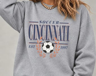 May include: A heather gray crewneck sweatshirt featuring a graphic print with the text SOCCER CINCINNATI EST 1997 in navy blue, accompanied by a black and white soccer ball illustration framed by orange laurel leaves on the front chest.