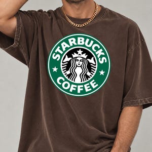 May include: A brown short-sleeve t-shirt featuring a large, circular green Starbucks Coffee logo printed on the chest. The logo includes the iconic siren graphic, white text, and two white stars on a green background. The shirt has a crew neckline.